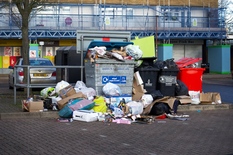 A collection of overflowing rubbish bins positioned on a paved sidewalk in an urban setting, with various types of waste including crumpled paper, cardboard boxes, plastic bags, and miscellaneous pack